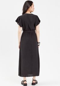 Robe maxi noire avec des manches courtes à volants, taille élastique et ourlet longueur cheville. Le mannequin porte des sandales noires à lanières et des bracelets.