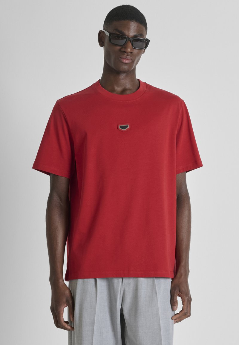 Hombre con gafas de sol negras, camiseta roja de manga corta con pequeño emblema en el pecho y pantalones anchos de pierna de color gris claro, de pie sobre un fondo liso.