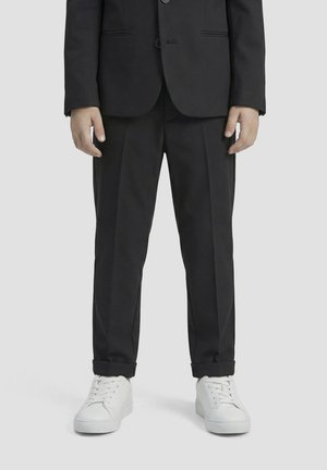 Persona con pantaloni da completo neri su misura con risvolti e sneakers bianche con lacci, in piedi davanti a uno sfondo chiaro e uniforme.