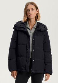 Čierna puff jacket s zipsom a zapínaním na patentky, veľká kapucňa a prešívaný dizajn. Nosená cez sivý top so zipsom a stojacím golierom.