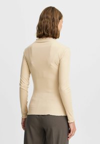 Beige gerippter Langarm-Oberteil mit einem Rüschensaum am Kragen und am Saum, taillierte Passform, von hinten vor einem hellen Hintergrund gezeigt.