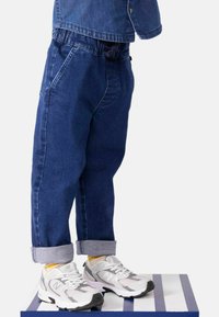 Bambino con pantaloni in denim blu arrotolati e sneakers bianco-grigie, in piedi su una piattaforma a strisce blu e bianche.