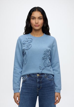 Frau mit langen dunklen Haaren, die einen blauen Sweatshirt mit strukturierten Blumenmustern und blaue Jeans trägt, vor einem einfarbigen Hintergrund.