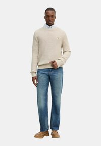 Maglione a coste beige, dal taglio rilassato, indossato sopra una camicia colletto azzurro; abbinato a jeans azzurri a gamba dritta e scarpe marroni.