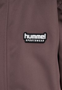 Brun sportsjakke med struktureret stof og et sort rektangulært mærke med "hummel" i hvid tekst og "SPORTSWEAR" under.