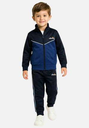 Jeune garçon souriant, portant un survêtement zippé bleu marine et bleu avec des bandes blanches et des baskets blanches, debout sur un fond blanc uni.