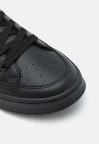 Sapatilha de couro preto com textura lisa, forma arredondada, bico perfurado e atacadores pretos contra um fundo branco.