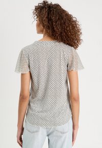 Blouse légère à motifs présentant des manches courtes évasées, un col rond, et une base crème avec des motifs ovale noirs. Coupe ample et décontractée.