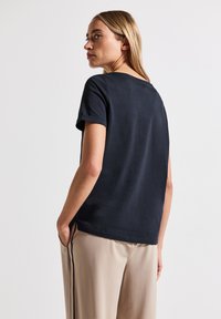 T-shirt bleu marine à manches courtes en coton doux, avec un col rond, des fentes sur les côtés et une coupe décontractée. Porté avec un pantalon beige clair.