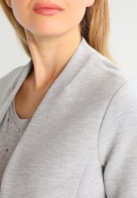 Close-up of a woman’s lower face and neck wearing a light gray blazer over a gray shirt with small heart patterns.