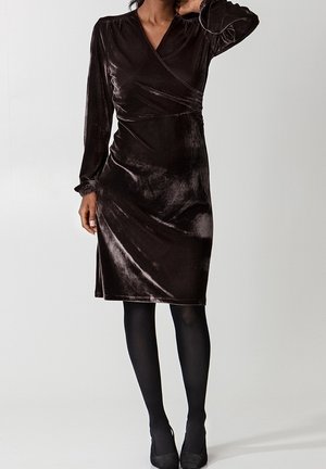 Kvinde iført en knælængde, langærmet mørkebrun fløjlsviklekjole, sorte strømpebukser og sorte hæle, stående med den ene hånd på halsen.