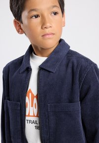 Laivastonsininen korjoreunatakki, jossa on etuvetoketju, kaksi rintataskua ja terävä kaulus, käytössä valkoisen graafisen t-paidan päällä.