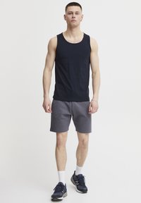 Schwarzes Tanktop mit rundem Ausschnitt, kombiniert mit grauen strukturierten Shorts. Das Modell trägt sportliche Schuhe und weiße Knöchelsocken. Einfaches, lässiges Outfit.