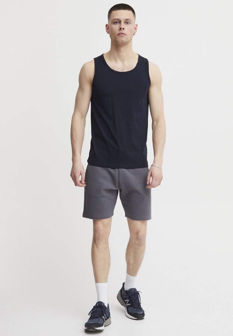 Schwarzes Tanktop mit rundem Ausschnitt, kombiniert mit grauen strukturierten Shorts. Das Modell trägt sportliche Schuhe und weiße Knöchelsocken. Einfaches, lässiges Outfit.