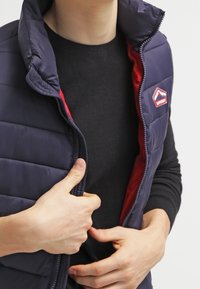Person, der eine marineblaue Steppweste mit rotem Futter über einem schwarzen Pullover trägt und den Reißverschluss der Weste mit beiden Händen schließt.
