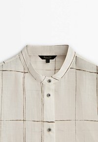 Beige button-up skjorte med stående krave, prydet med et rutemønster i brun, lavet af letvægtsstof, med synlige knapper og syninger.