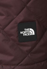 Bordeaux prošívaná látka s černou čtvercovou nálepkou s bílým textem a zakřiveným logem, na kterém je napsáno "THE NORTH FACE."