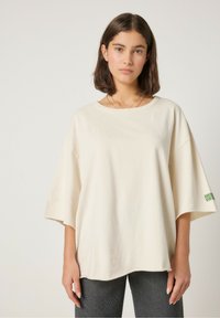 Kremowy, oversize'owy t-shirt z krótkimi rękawami i okrągłym dekoltem. Na rękawie znajduje się zielony nadruk. Wykonany z bawełny o gładkiej fakturze.