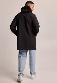 Chaqueta negra con capucha y corte holgado, con un largo posterior, fabricada en un material suave, combinada con jeans de mezclilla azul claro y zapatillas blancas.