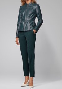 Teal leather peplum jacket with a zip front, paired with fitted dark green trousers and white high-heeled shoes. Smooth texture, structured design.