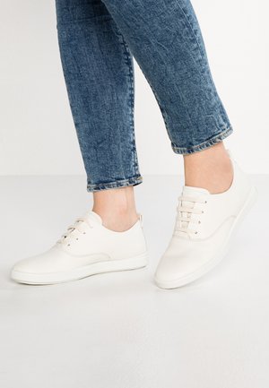 Osoba nosząca białe trampki na sznurowadła i niebieskie dżinsy, stojąca na białej podłodze na jednolitym białym tle.