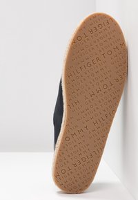 Semelle de chaussure avec motif répétitif de texte "Tommy Hilfiger" en caoutchouc brun, affichant un marquage texturé sur une tige noire.