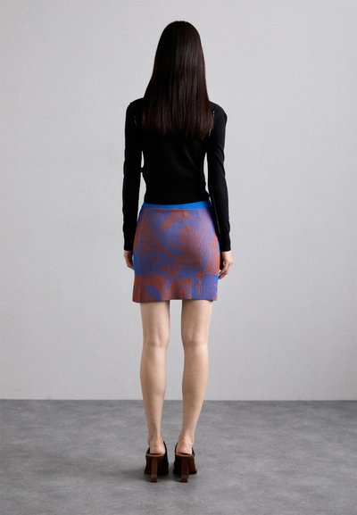 Bluză neagră cu mânecă lungă, fustă mini cu model albastru și portocaliu, și pantofi cu toc pătrat de culoare maro. Textură netedă, design ajustat și culori contrastante.