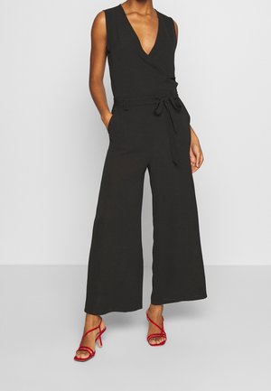 Kobieta w czarnym, bezrękawnikowym kombinezonie z szerokimi nogawkami i wiązanym paskiem w talii, w połączeniu z czerwonymi sandałami na wysokim obcasie z paskami.