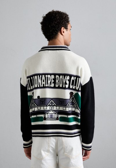 Un pull en tricot crème et noir arborant un motif de maison avec des accents verts et violets. Poignets et col côtelés, coupe décontractée.