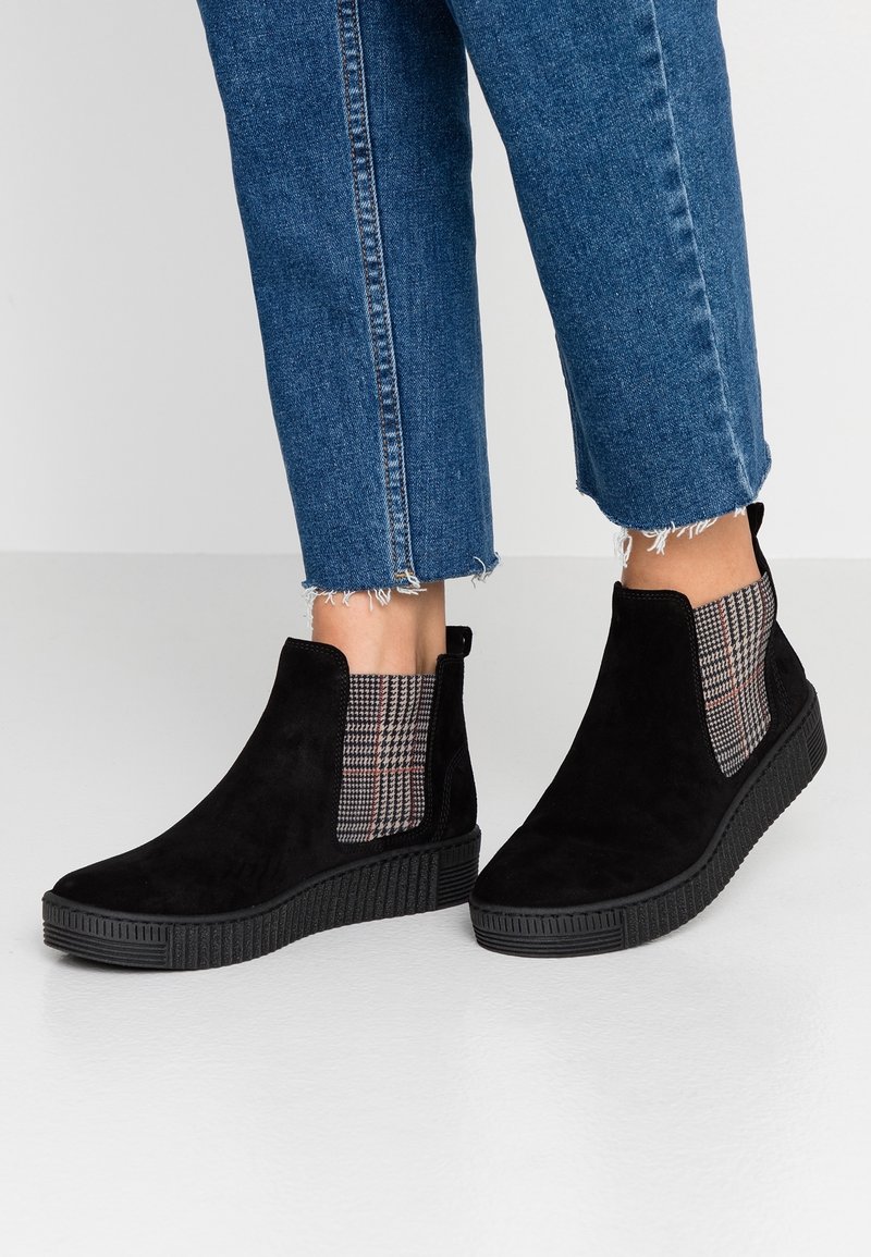 Femme portant des bottines en daim noir avec des empiècements latéraux élastiques à carreaux et des semelles en caoutchouc noir, assorties à un jean court bleu.