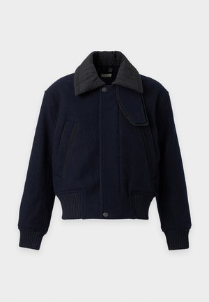 Homme portant une veste bleu foncé, un pull bleu, un jean large délavé et des chaussures noires, debout devant un fond clair uni.