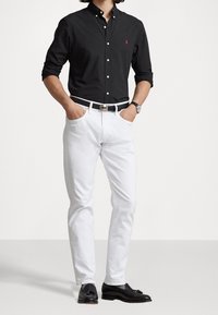 Camicia nera a maniche lunghe con logo rosso, jeans bianchi, cintura nera e mocassini neri. I jeans hanno un taglio slim.