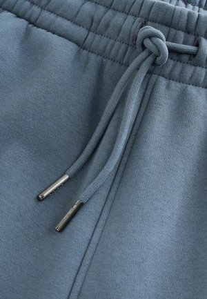 A világoskék melegítőnadrág texturált anyagból készült, rugalmas derékpántja állítható húzózsinórokkal rendelkezik, melyek fémes végződéssel vannak ellátva.