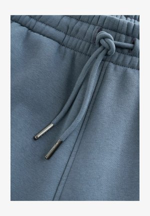 A világoskék melegítőnadrág texturált anyagból készült, rugalmas derékpántja állítható húzózsinórokkal rendelkezik, melyek fémes végződéssel vannak ellátva.