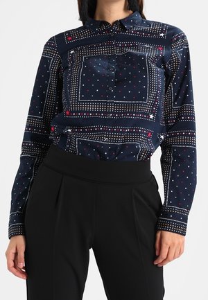 Granatowa koszula na guziki z białymi i czerwonymi wzorami gwiazdek i kropek, zestawiona z czarnymi, wysmuklonymi spodniami z wysokim stanem.