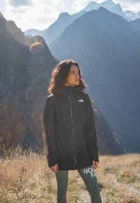 Giacca nera impermeabile con colletto alto, zip frontale e logo. Indossata sopra leggings verdi aderenti con la scritta "NORTH FACE". Sfondo montano.