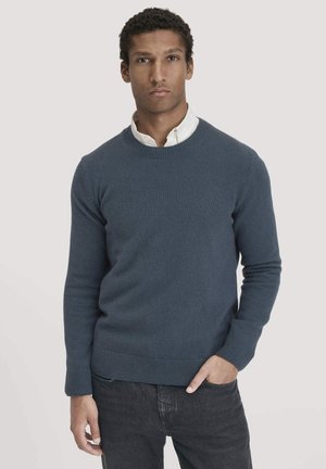 Dunkelblaues gestricktes Sweatshirt mit Rundhalsausschnitt, gerippten Bündchen und Saum. Über einem weißen Hemd mit Kragen getragen, kombiniert mit dunklen Jeans.