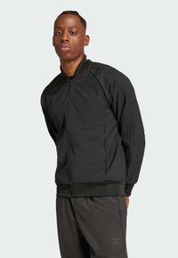 Čierna bundu na zips s vysokým golierom, vyrobenú z textúrovaného materiálu, s bočnými vreckami a tromi vertikálnymi pásmi na každom rukáve.