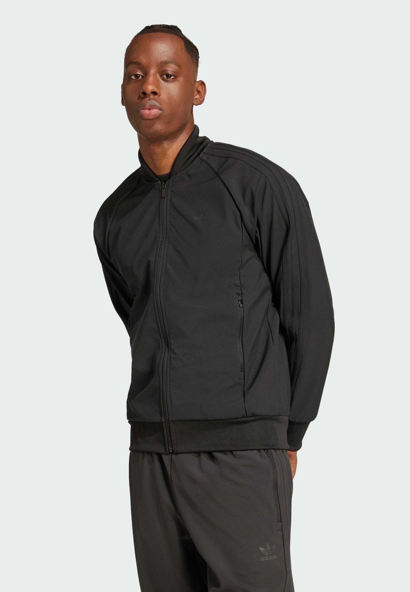 Čierna bundu na zips s vysokým golierom, vyrobenú z textúrovaného materiálu, s bočnými vreckami a tromi vertikálnymi pásmi na každom rukáve.