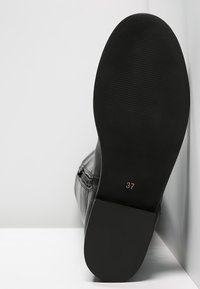 Bottine en cuir noir, semelle tournée vers l'avant, taille 37 marquée au bas, positionnée contre un fond blanc.