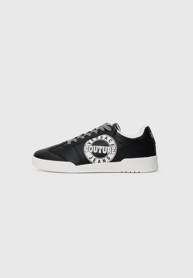 Sapatilhas baixas pretas com sola branca, atacadores pretos e brancos com padrão, e o logo "Versace Jeans Couture" na lateral.