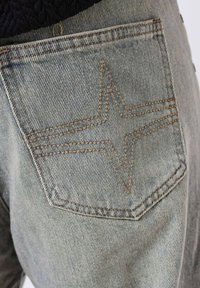 Bolsillo de vaqueros de mezclilla con un diseño de puntada en forma de estrella en tela azul claro, con hilo marrón en contraste y una superficie texturizada.