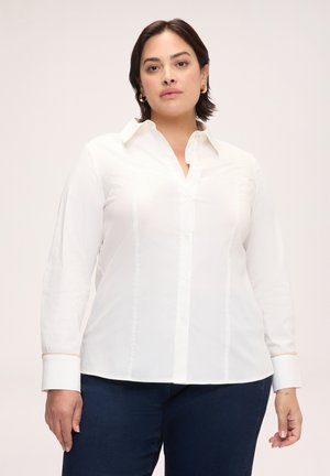 Frau trägt ein weißes Hemd mit goldumrandeten Manschetten und dunkelblaue Jeans vor einem schlichten hellen Hintergrund.