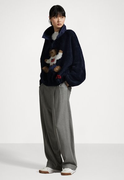 Marineblauer Fleece-Pullover mit Teddy-Bären-Design, hohem Kragen und roten Akzenten. Kombiniert mit grauen weit geschnittenen Hosen und weißen Sneakers.