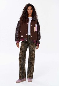 Brązowa kurtka varsity ze skórzanymi rękawami, różowymi akcentami i naszywkami, w połączeniu z spodniami w leopardzi wzór w stylu dzwonów oraz fioletowymi sneakersami.