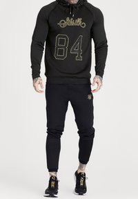 Felpa nera con logo dorato "SikSilk" e "84" sul davanti, abbinata a pantaloni jogger neri a gamba affusolata con un piccolo logo. Polsini a costine e cappuccio con cordino.
