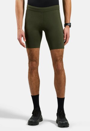 Man wearing olive green fitted athletic shorts, black socks, black sneakers, and a black watch, standing against a plain background.