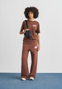 Καφέ βαμβακερό oversized μπλουζάκι με ροζ λογότυπο, σετ με ταιριαστά παντελόνια φαρδιάς γραμμής. Μαύρη τσάντα με χρυσή λεπτομέρεια. Αθλητικά παπούτσια ολοκληρώνουν την εμφάνιση.