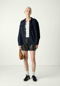 Giacca di jeans scuri con chiusura a bottoni, abbinata a pantaloni corti in pelle nera. Gli accessori includono una borsa marrone e sneakers nere con accenti bianchi.