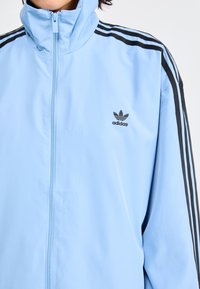 Chubasquero azul claro con cremallera, hecho de una tela suave, que presenta acentos de tres rayas negras en los hombros y un logo de Adidas bordado en negro.